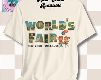 May include: Cream-colored t-shirt featuring the text "WORLD'S FAIR NEW YORK 1964-1965" in a colorful, vintage design. The graphic includes illustrations of buildings and children with balloons. The words "More Colors Available" are also printed on the shirt.