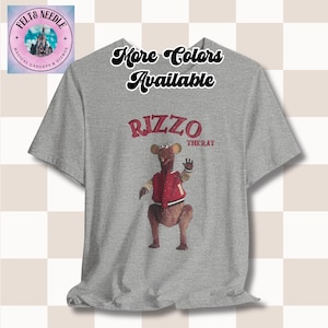 May include: Grey t-shirt featuring a graphic of Rizzo the Rat from the Muppets. The graphic depicts Rizzo wearing a red jacket and standing with his arms outstretched. The text "Rizzo the Rat" is printed below the graphic.
