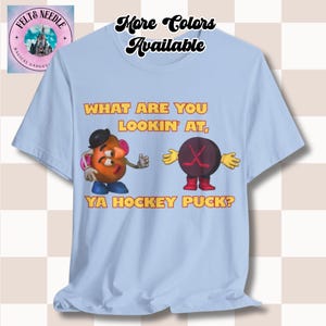 May include: Pale blue t-shirt with a cartoon graphic and the text "WHAT ARE YOU LOOKIN' AT, YA HOCKEY PUCK?". The shirt also has the text "More Colors Available" at the top. The shirt is a light blue colour.