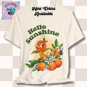Puede incluir: Una camiseta blanca con un pájaro naranja de dibujos animados y flores de azahar. El texto "Hello Sunshine" está impreso en la camiseta.