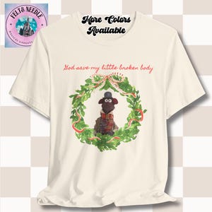 May include: Cream-colored t-shirt featuring a cartoon sheep character wearing a hat and scarf, inside a green wreath with red ribbons. Text reads "God save my little broken body" and "More Colors Available".