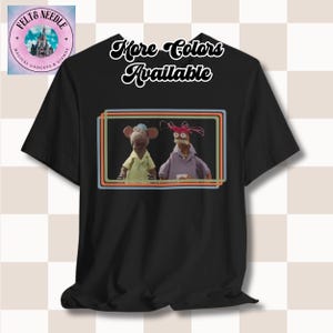 May include: Black t-shirt featuring a framed image of two Muppet characters. The frame has orange, blue, and yellow lines. The text "More Colors Available" is above the frame. A logo is in the upper left corner.
