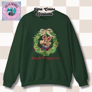 Könnte beinhalten: Ein dunkelgrünes Sweatshirt mit einer Cartoon-Kaninchenfigur in einem Weihnachtskranz. Der Kranz hat rote und weiße Akzente und eine Schleife oben. Der Text "Penny for the song, guwhan?" steht unten.