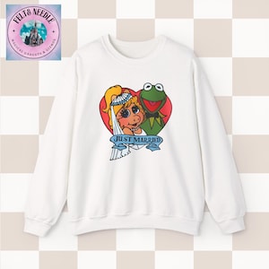 May include: A white crewneck sweatshirt featuring a graphic of Miss Piggy and Kermit the Frog inside a red heart. Miss Piggy wears a white bridal veil and a pearl necklace. Below them is a blue banner with the text JUST MARRIED in black letters.