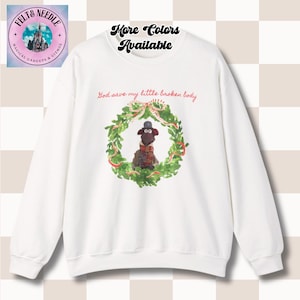 May include: White sweatshirt with a graphic of a cartoon character inside a green wreath, with the text "God save my little broken body." The words "More Colors Available" are above the graphic. A logo is in the upper left corner.
