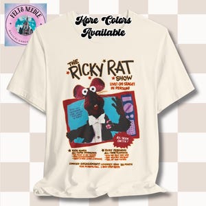 May include: Off-white t-shirt with a graphic promoting "The Ricky Rat Show." The design shows a cartoon rat in a tuxedo, the show's title, and the words "Live! On Stage! In Person!" The shirt also has the text "More Colors Available."