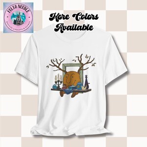 May include: White t-shirt with a cartoon illustration of a brown, smiling face with antlers, sitting at a table with candles, teacups, and a pipe. The text "More Colors Available" is at the top of the image.