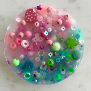 May include: A clear round resin coaster featuring a mix of pink, green and blue beads, sequins and other small decorative elements. A pink wooden heart with white polka dots is embedded in the resin.