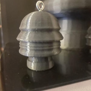 Könnte beinhalten: Ein dunkelgrauer, 3D-gedruckter Ornament mit einem geschichteten, glockenförmigen Design. Es hat eine kleine, silberne Metallschlaufe oben zum Aufhängen. Das Ornament wird auf einer reflektierenden schwarzen Oberfläche präsentiert.