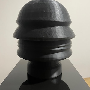 Könnte beinhalten: Eine schwarze, geschichtete Skulptur mit abgerundeter Oberseite, montiert auf einem schwarzen Sockel. Die Skulptur hat ein einzigartiges, gestapeltes Design. Der Sockel hat italienischen Text. Es ist ein dekoratives Kunstwerk.