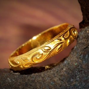 May include: A gold ring featuring a textured surface with a raised, polished vine and leaf pattern that wraps around the entire band. The metal has a bright, reflective finish on the raised elements, contrasting with the matte, granular texture of the background.