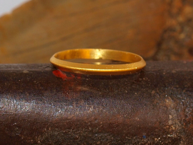 May include: A simple gold band ring, with a smooth, polished finish. The ring is sitting on a dark, textured surface.