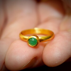 Puede incluir: Un anillo de oro con una gema verde redonda y brillante. El anillo tiene un dise&ntilde;o cl&aacute;sico, con una banda lisa y pulida. La gema es el punto focal, engastada en un bisel. El anillo se sostiene en una mano.