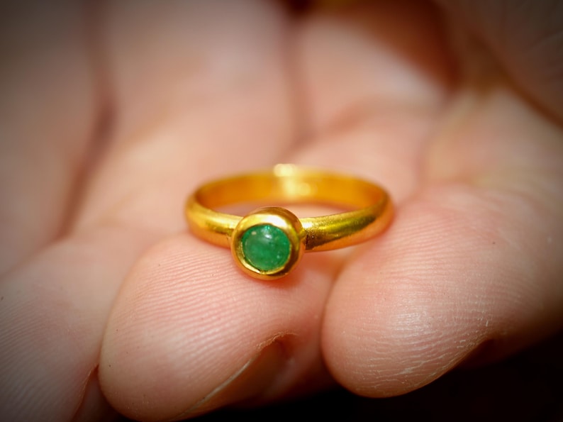 Puede incluir: Un anillo de oro con una gema verde redonda y brillante. El anillo tiene un dise&ntilde;o cl&aacute;sico, con una banda lisa y pulida. La gema es el punto focal, engastada en un bisel. El anillo se sostiene en una mano.