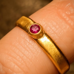 May include: A gold ring with a bright red gemstone set in the center. The ring has a smooth, polished band and a bezel setting that holds the gemstone securely. The ring is shown on a finger.