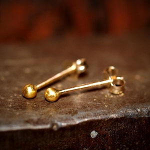 May include: Two small, gold-colored stud earrings with a ball at the end of each post. The earrings are displayed on a dark, textured surface, with a blurred brown background.