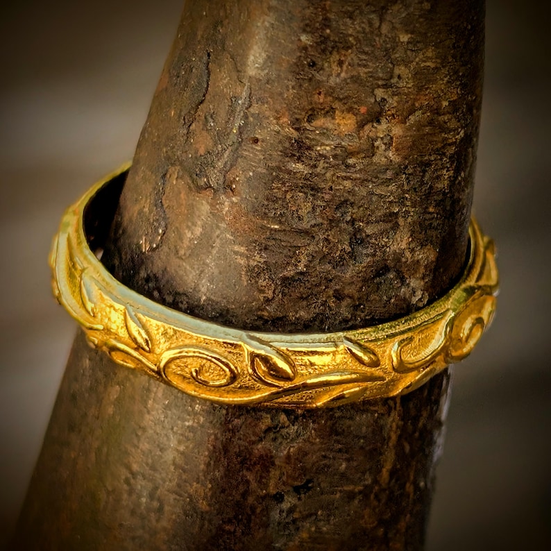May include: A gold ring featuring a continuous embossed pattern of swirling vines and leaves. The band has a polished finish and is positioned around a dark, textured, conical object that serves as a display stand for the jewelry piece.