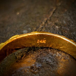 May include: A gold ring with a smooth, polished surface and a rounded band. The interior of the band is clearly stamped with the text 24K, indicating the purity of the gold. The metal has a bright, reflective yellow tone throughout the piece.