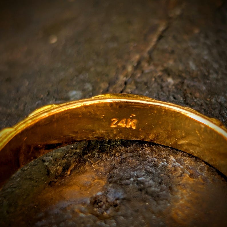 May include: A gold ring with a smooth, polished surface and a rounded band. The interior of the band is clearly stamped with the text 24K, indicating the purity of the gold. The metal has a bright, reflective yellow tone throughout the piece.