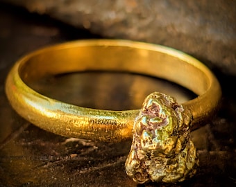May include: A gold ring featuring a prominent, raw, textured gold nugget attached to the side of a smooth, polished, circular band. The metal has a bright, reflective yellow finish with visible variations in the surface texture of the nugget.
