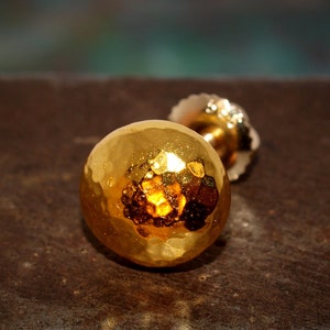 May include: A gold-toned earring with a hammered, round ball design. The earring is on a brown surface.