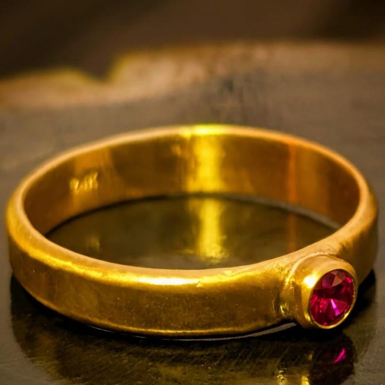 May include: A gold ring with a round, polished band and a single, round, red gemstone. The ring is set in a simple bezel setting, and the gold has a warm, slightly aged appearance. The ring is a classic piece of jewelry.