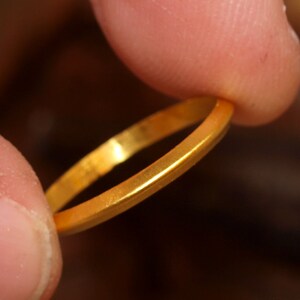 May include: A simple gold band ring, held between two fingers.