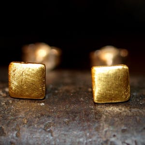 May include: Close-up of two small, square gold stud earrings. The earrings have a textured surface and are set against a dark background. The earrings are approximately 0.25 inches in size.