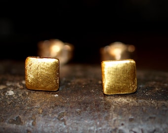 May include: Close-up of two small, square gold stud earrings. The earrings have a textured surface and are set against a dark background. The earrings are approximately 0.25 inches in size.