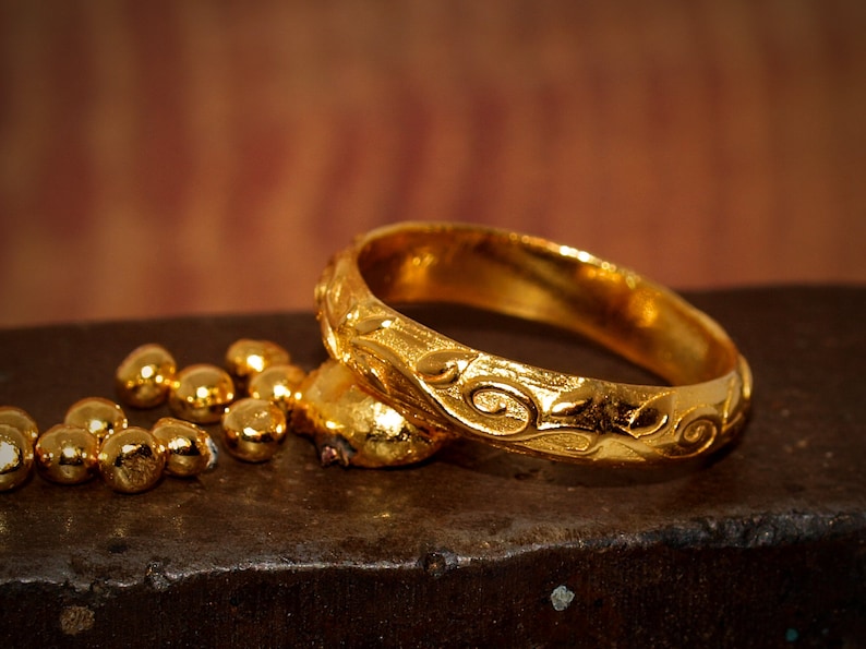 May include: A polished gold ring featuring an embossed swirling vine pattern along its outer band, accompanied by several small, rounded gold nuggets resting nearby, showcasing a bright, reflective metallic finish and intricate relief detailing.