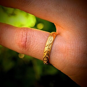 May include: A gold-toned ring featuring an embossed, continuous swirling vine and leaf pattern around the entire band. The metal surface has a polished finish that catches the light, highlighting the intricate, raised botanical engravings.
