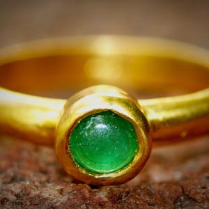 Puede incluir: Un anillo de oro con una piedra preciosa verde brillante en el centro. La banda del anillo es de oro pulido y la piedra preciosa est&aacute; engastada en un bisel de oro. El anillo se muestra sobre una superficie marr&oacute;n texturizada.