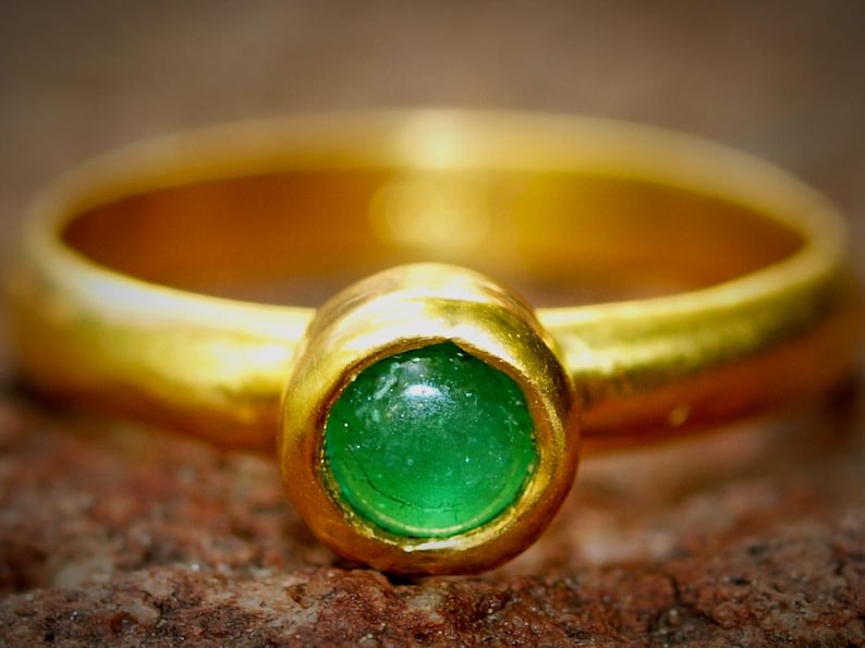 Puede incluir: Un anillo de oro con una piedra preciosa verde brillante en el centro. La banda del anillo es de oro pulido y la piedra preciosa est&aacute; engastada en un bisel de oro. El anillo se muestra sobre una superficie marr&oacute;n texturizada.