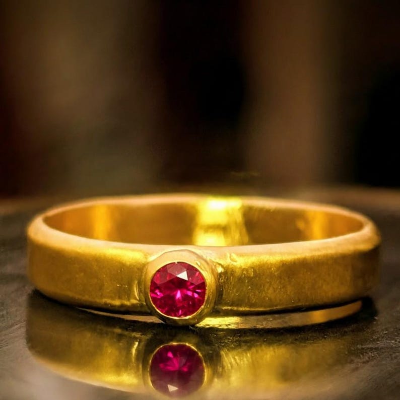 May include: A gold ring with a round, bright red gemstone set in the center. The ring has a simple, classic design with a polished finish. The gemstone is the focal point, catching the light and reflecting a vibrant color. The ring is a piece of jewelry.