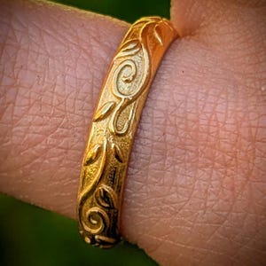 May include: A gold ring featuring a textured surface with an embossed pattern of swirling vines and leaves. The metal has a matte finish, and the design wraps around the band, showcasing intricate, raised organic shapes across the entire visible exterior.