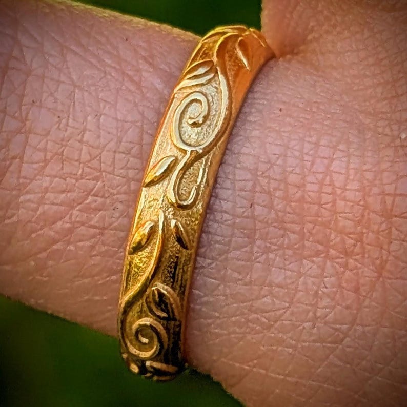 May include: A gold ring featuring a textured surface with an embossed pattern of swirling vines and leaves. The metal has a matte finish, and the design wraps around the band, showcasing intricate, raised organic shapes across the entire visible exterior.