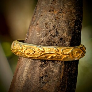 May include: A gold ring featuring a continuous embossed vine and leaf pattern around the entire band. The metal surface has a textured, matte finish with raised, polished details that catch the light, creating a contrast between the gold elements.