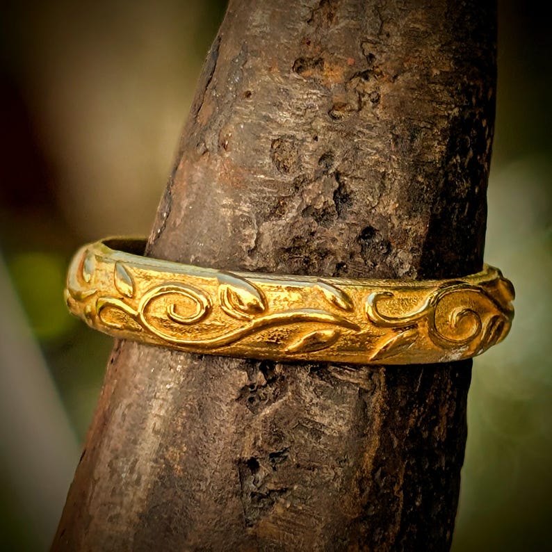 May include: A gold ring featuring a continuous embossed vine and leaf pattern around the entire band. The metal surface has a textured, matte finish with raised, polished details that catch the light, creating a contrast between the gold elements.
