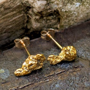 May include: A pair of gold stud earrings featuring irregular, textured nugget-shaped designs. Each earring has a polished gold post and a secure butterfly back, showcasing a raw, organic appearance with a bright, reflective metallic finish.