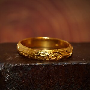 May include: A gold ring featuring a continuous embossed scrollwork pattern around the entire band. The metal has a textured, matte finish with polished highlights on the raised swirling designs, creating a distinct contrast across the surface.