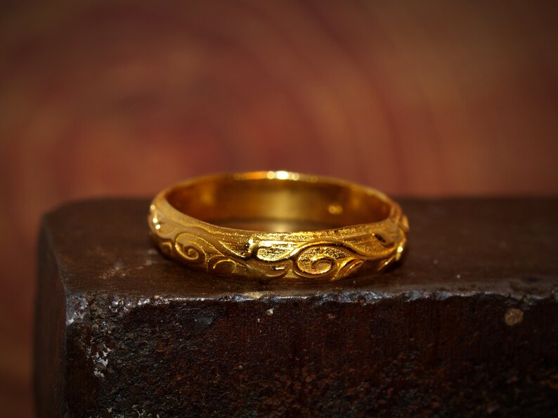 May include: A gold ring featuring a continuous embossed scrollwork pattern around the entire band. The metal has a textured, matte finish with polished highlights on the raised swirling designs, creating a distinct contrast across the surface.