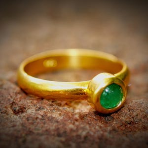 Puede incluir: Un anillo de oro con una piedra preciosa verde brillante y redonda. El anillo tiene una banda lisa y pulida y un engaste de bisel para la esmeralda. El anillo est&aacute; sobre una superficie marr&oacute;n texturizada.