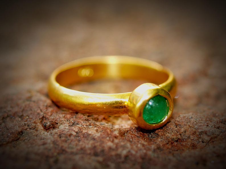 Puede incluir: Un anillo de oro con una piedra preciosa verde brillante y redonda. El anillo tiene una banda lisa y pulida y un engaste de bisel para la esmeralda. El anillo est&aacute; sobre una superficie marr&oacute;n texturizada.