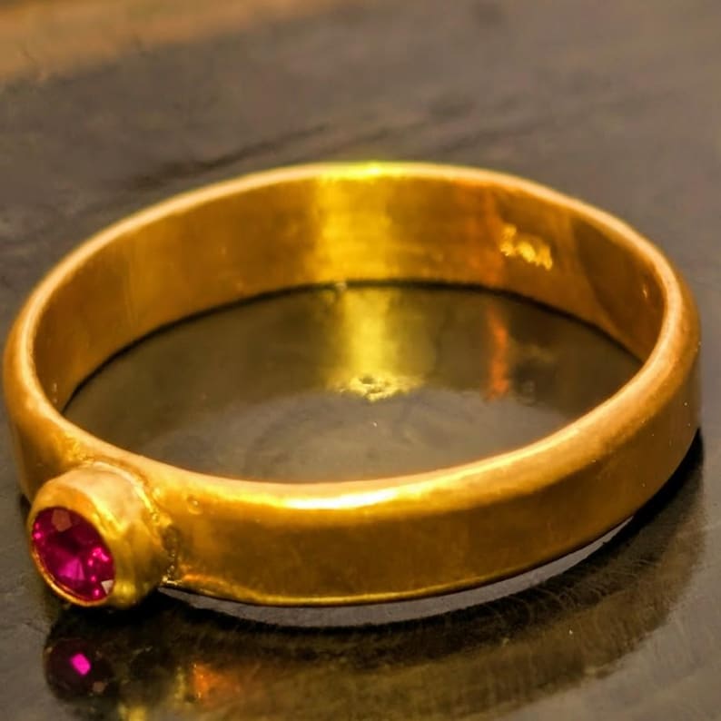 May include: A gold ring with a single, round, red gemstone. The ring has a smooth, polished surface and a classic design. The gemstone is set in a simple bezel setting, adding a touch of color and sparkle to the ring.