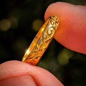 May include: A gold ring featuring a continuous embossed pattern of swirling vines and leaves. The metal surface has a polished finish that catches the light, highlighting the intricate relief details carved into the band's exterior circumference.