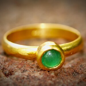 Puede incluir: Un anillo de oro con una piedra preciosa verde redonda y brillante en el centro. El anillo tiene un dise&ntilde;o simple y cl&aacute;sico con una banda lisa y pulida. La piedra preciosa es de un color verde vibrante.
