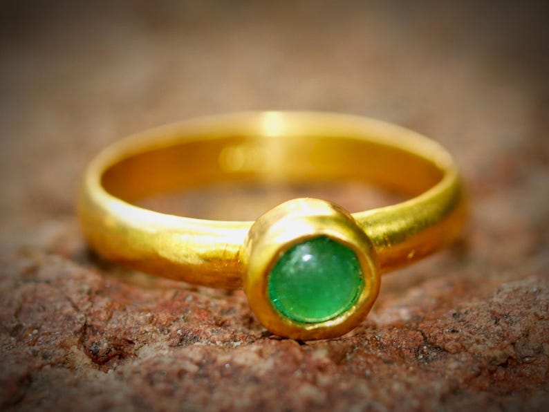 Puede incluir: Un anillo de oro con una piedra preciosa verde redonda y brillante en el centro. El anillo tiene un dise&ntilde;o simple y cl&aacute;sico con una banda lisa y pulida. La piedra preciosa es de un color verde vibrante.