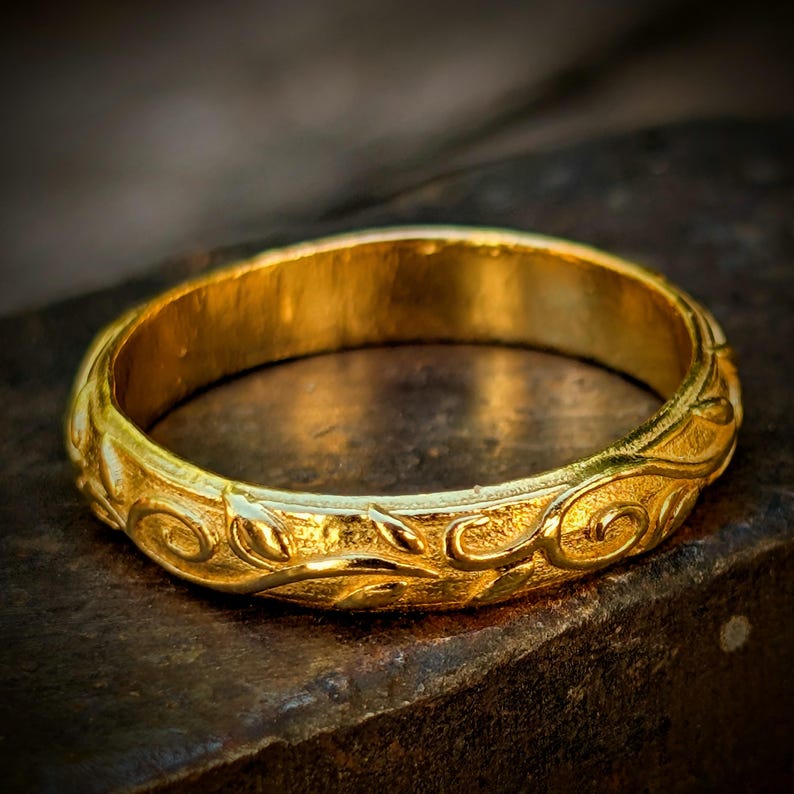 May include: A gold band ring featuring a continuous embossed pattern of swirling vines and leaves. The metal surface has a textured finish, and the design wraps entirely around the circumference of the circular band, creating a detailed relief.
