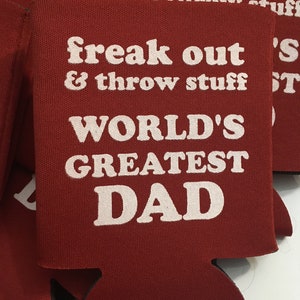 May include: Red can cooler with white text that reads "freak out & throw stuff WORLD'S GREATEST DAD".