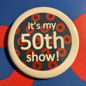 May include: A round button with a white border and the text "It's my 50th show!" in red and white. The button's background is navy blue with red circles. The background is blue with red circles.